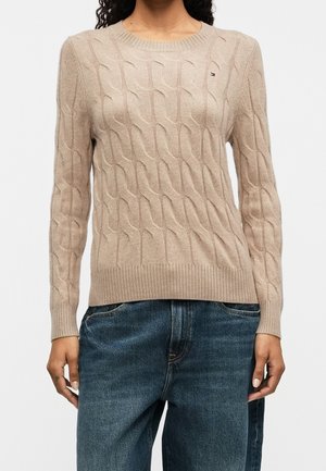 Kobieta ubrana w beżowy sweter o splocie warkoczowym z okrągłym dekoltem i niebieskie dżinsy, stojąca na tle jasnego, jednolitego tła.