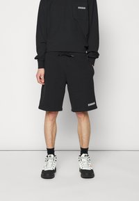 Un sweat-shirt en coton noir avec une poche poitrine et un short noir à cordon de serrage. Détail de logo notable à l'ourlet et associé à des baskets noir et blanc.