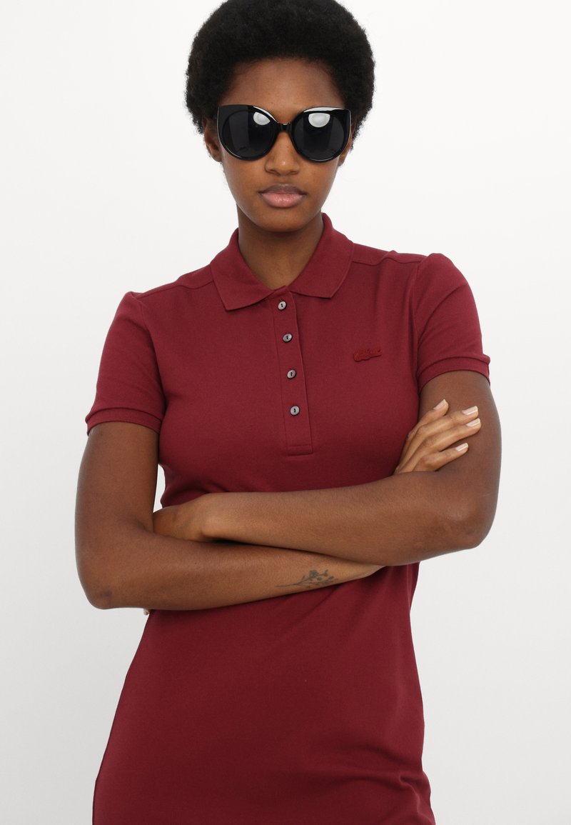 Polo shirt bourgogne avec un col et une patte de boutonnage à trois boutons, ornée d'un petit logo brodé. Le modèle porte des lunettes de soleil noires surdimensionnées.