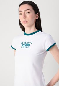 White fitted t-shirt with teal ribbed collar and sleeves, featuring a bold teal graphic of "CALVIN KLEIN 1968" at the center.