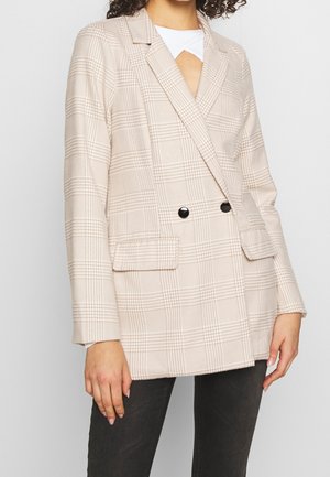 Blazer de cuadros beige de doble botonadura con botones negros, usado sobre una blusa blanca, combinado con pantalones oscuros, mostrado desde los hombros hasta las caderas.