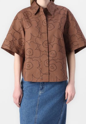 Chemise marron à manches courtes avec boutons et grandes fleurs noires en contour, portée avec une jupe en jean bleue.