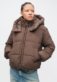 Jonge persoon met gevlochten haar, gekleed in een bruine pufferjas met de handen in de zakken, kijkt naar de zijkant, tegen een effen witte achtergrond.