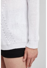 Biały dziergany sweter o luźnym splotzie, z prążkowanymi mankietami i dołem. Noszony na czarnych szortach, podkreślający fakturę i lekkość.