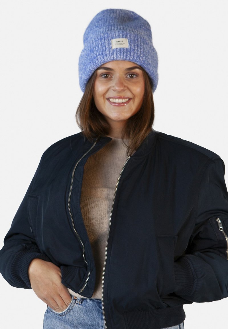 Navy bomber jacket with a ribbed collar, a beige sweater underneath, and a textured light blue knit beanie with a tag.