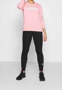Felpa Adidas rosa con logo bianco, abbinata a leggings neri aderenti con strisce laterali bianche e scarpe da ginnastica nere.