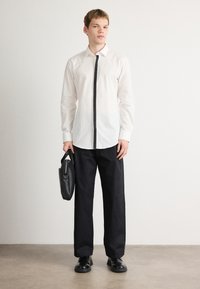 Witte blouse met lange mouwen en zwarte middenknopen, gecombineerd met zwarte broek. Het model houdt een zwarte leren tas vast en draagt zwarte schoenen.