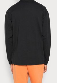 Person wearing a black long-sleeve shirt and orange pants, shown from the back against a plain light background.