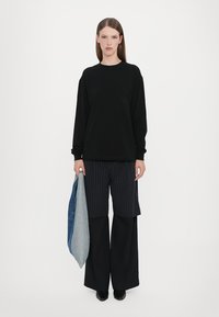Svart långärmad sweatshirt med ficka; ihop med randiga vida byxor. Håller ett vikt jeansplagg i ena handen.