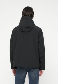 Mężczyzna widziany z tyłu, ubrany w czarną kurtkę z kapturem i niebieskie dżinsy na jednolitym białym tle.