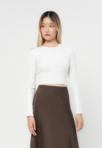 Bijela dugorukava crop top majica s rebrastom teksturom, u kombinaciji s smešnim suknjom koja ima glatku površinu i bočni prorez. Jednostavan i prilagođen dizajn.