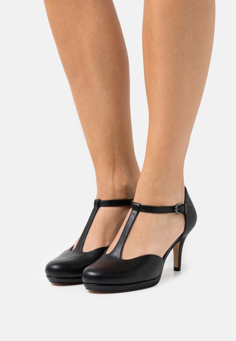 Talons hauts en cuir noir avec une bride en T et un bout rond, sangle de cheville avec boucle en argent. Texture lisse et design épuré.