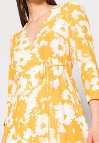 Robe florale jaune avec des motifs blancs, décolleté en V profond, manches trois-quarts et une ceinture à nouer à la taille, fabriquée en tissu doux.