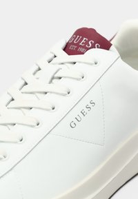 Baskets blanches Guess avec lacets, étiquette rouge sur la languette indiquant "GUESS EST. 1981" et discret logo de la marque sur le panneau latéral.