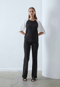 Haut noir et blanc avec des manches évasées, coupe slim, associé à un jean noir évasé et des sandales noires. Fond uni.