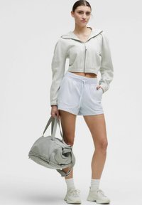 Sudadera corta con cremallera en gris claro y pantalones cortos con cinturilla elástica blanca, combinados con un bolso de lona gris. El modelo lleva calcetines blancos y zapatillas deportivas.