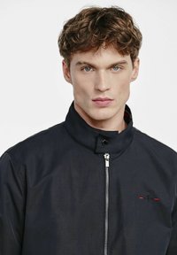 Niebieska lekka kurtka z wysokim kołnierzem, zamek błyskawiczny z przodu i czerwonym akcentem. Gładka tekstura, casualowy design odpowiedni na różne warunki pogodowe.