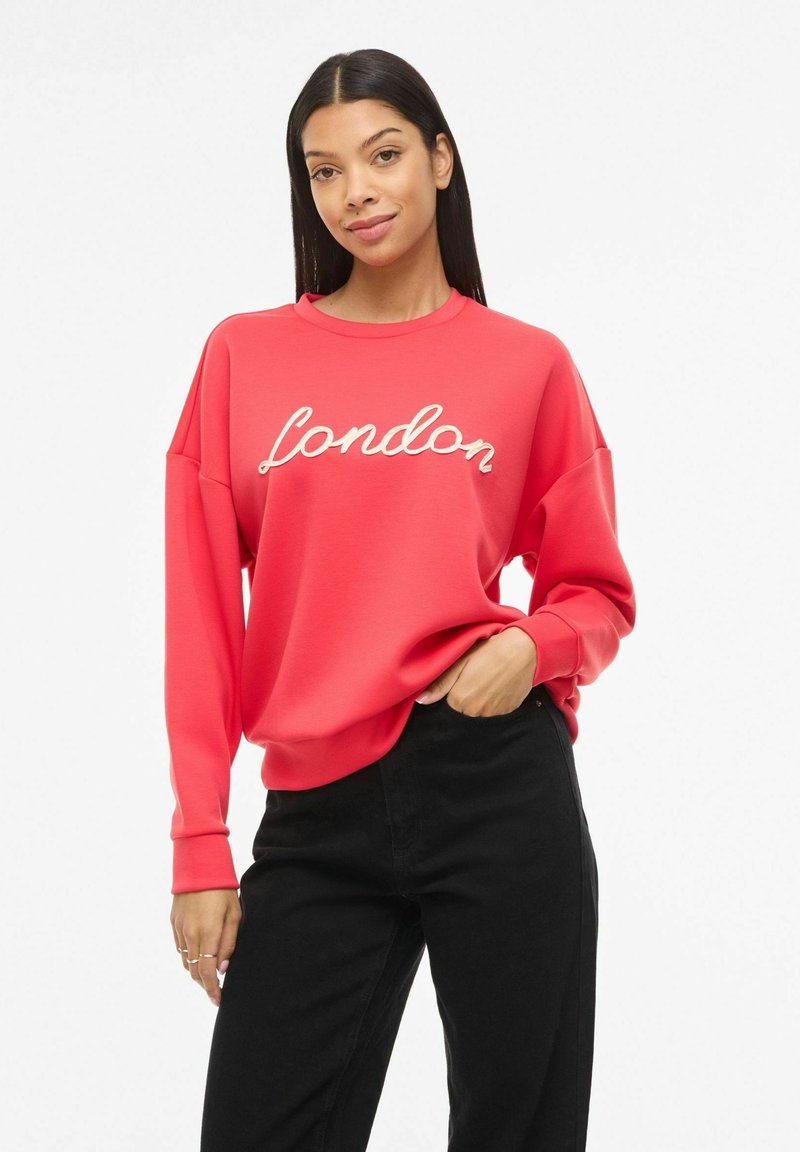 Jeune femme aux longs cheveux noirs portant un sweat-shirt rouge avec le texte « London » et un pantalon noir, debout avec une main dans la poche.