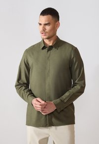 Camisa verde de manga larga en una tela suave y texturizada con cuello puntiagudo y cierre frontal de botones. Modelo posa con las manos entrelazadas.