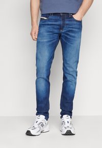 Jeans de mezclilla azul con un corte ajustado, que presentan costuras en contraste, cinco bolsillos y un cierre con cremallera clásico. Combinados con zapatillas deportivas blancas.