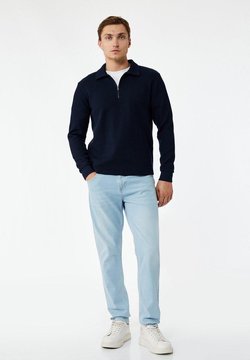 Ung mand, der står, iført en navyblå pullover med lynlås foran, lyseblå jeans og hvide sneakers på en hvid baggrund.