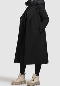 Personne vêtue d'un long manteau noir et de leggings noirs avec des bottines montantes beiges épaisses à lacets, debout devant un fond neutre.