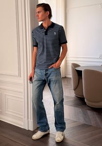 Jeune homme portant un polo rayé bleu et noir, un jean bleu clair et des baskets blanches, debout à l'intérieur près d'une table à manger et des chaises.