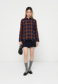 Woman wearing a brown and blue plaid shirt, navy mini skirt, gray knee-high socks, black flats, holding a black handbag, standing against white background.