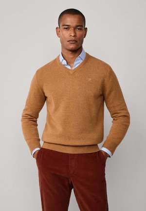 Suéter de color camel con escote en V, hecho de una tela suave, combinado con una camisa de cuello clara de color azul debajo. Textura lisa con puños acanalados.