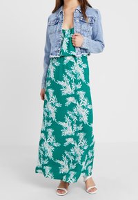 Jeansjacka med en figurnära design, ljusblå färg. Grön maxiklänning med vita bladmönster, mjukt tyg och justerbara axelremmar. Vita sandaler.