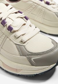 Svetlé krémové bežecké topánky s textúrovaným sieťovaným zvrškom, šedými akcentmi a fialovými šnúrkami. Majú polstrovanú podrážku a výrazné šitie.