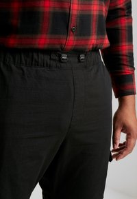 Schwarze Hose mit elastischem Bund und Kordelzug, kombiniert mit einem rot-schwarz karierten Hemd mit Knopfdetails. Strukturierter Stoff ist sichtbar.
