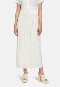 Maxi-Kleid aus weichem weißen Stoff mit Raffung in der Taille und Kordelzugdetails, entspannt geschnitten und mit kurzen Ärmeln. Getragen mit beigen Plateauschuhen.