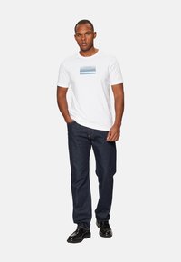 Camiseta blanca de algodón con diseño gráfico azul, combinada con jeans de mezclilla oscura y zapatos negros, con un corte clásico y escote redondo.