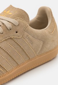 Béžová semišová teniska s gumovou podrážkou, třemi vodorovnými proužky a reliéfním nápisem "SAMBA". Design doplňují tkaničky a měkký límec.