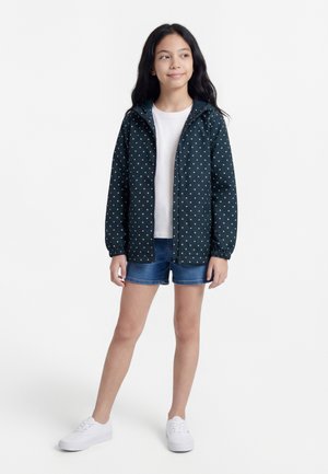 Dievča v tmavomodrej kapucňovej bunde s bodkami, bielom tričku, rifľových šortkách a bielych teniskách stojace na bielom pozadí.