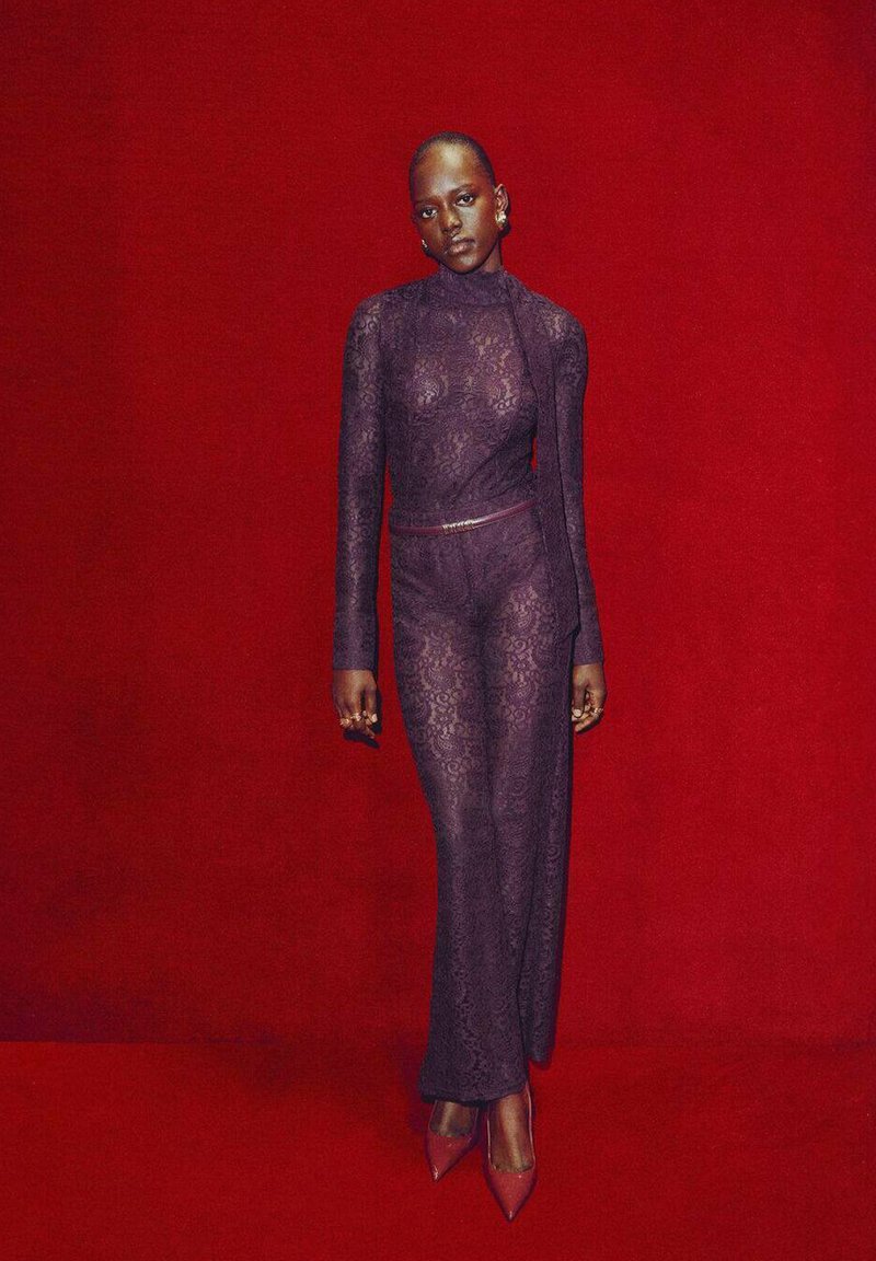 Mono de encaje morado con mangas largas, que presenta un cuello alto y una abertura lateral, complementado con un cinturón delgado y tacones de punta a juego.