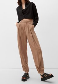 Zwarte cropped shirt met knopen, gecombineerd met tan geplooide broeken met een elastische tailleband en zwarte plateausandalen.