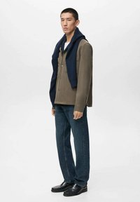 Chaqueta de gran tamaño en color oliva oscuro con un suéter de punto azul marino caído sobre los hombros, combinado con jeans de pierna recta y zapatos formales negros.