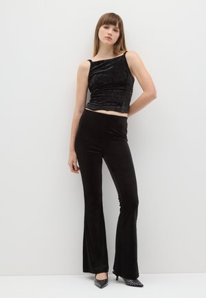 Top corto in velluto nero con spalline sottili abbinato a pantaloni neri a zampa. L'outfit presenta una texture elegante e una silhouette aderente.