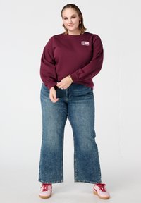Bordeauxrode sweatshirt met pofmouwen, ronde hals en een logo aan de voorkant. Gecombineerd met lichtblauwe wijde jeans en roze sneakers met rode veters.
