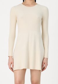 Robe en maille crème avec manches longues, texture côtelée, encolure ronde et ourlet légèrement plus long à l'arrière. Tissu doux avec une silhouette ajustée.