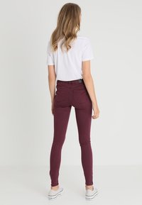 Jeans skinny burdeos de cintura alta, con una textura suave y bolsillos traseros, combinados con una camiseta blanca de manga corta y zapatillas.