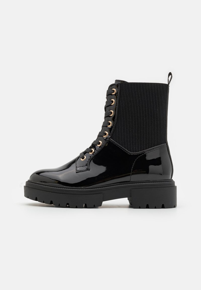 Botte de combat noire en cuir verni avec cheville tricotée côtelée, œillets dorés, semelle épaisse à crampons et languette à l'arrière, vue de côté.