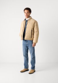 Geacă puffer bej cu guler înalt, cu un buzunar cu fermoar, purtată peste o cămașă din denim și asortată cu blugi albaștri și cizme bej.