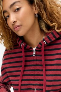 Νεαρή γυναίκα με σγουρά μαλλιά που φοράει ασημένια κρίκους σκουλαρίκια και φούτερ με φερμουάρ με κόκκινες και μαύρες ρίγες και κορδόνια.
