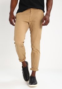 Khaki chinos i slät bomull med smal passform, upprullade fållar och sidofickor, i kombination med svarta sneakers.