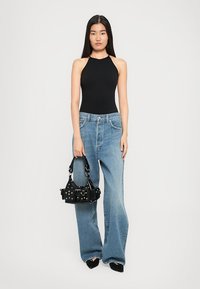 Femme aux longs cheveux noirs portant un haut noir sans manches et un jean bleu à jambes larges, tenant un sac à main noir clouté.