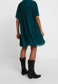 Teal pleated dress with short sleeves, flowing design, and translucent hem, paired with black leather cowboy boots, featuring curved tops.