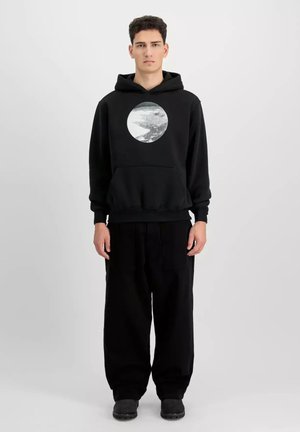 Young man standing, wearing black hoodie with circular grayscale graphic, loose black pants, and black shoes, neutral gray background.